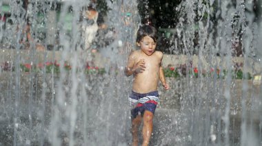 Küçük çocuk yaz günü şehir parkında su jetlerinde koşuyor. Küçük çocuk ferahlatıcı suda koşuyor yavaş çekimde sıçrıyor