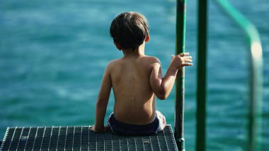 One pensive child seated by pier looking at water landscape. Thoughtful wet kid daydreaming at shore. Contemplative childhood emotion