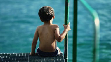 One pensive child seated by pier looking at water landscape. Thoughtful wet kid daydreaming at shore. Contemplative childhood emotion
