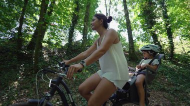 Anne bisiklete biniyor ve çocuğu arka koltukta oturuyor. Orman yolunda.