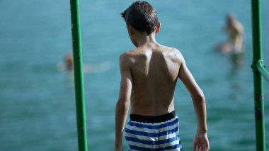 Düşünceli küçük bir çocuk göl kıyısında ayakta duruyor. Yürüyor ve mayoyu kısa sürede ayarlıyor. Islak, düşünceli, ciddi bir erkek çocuk.