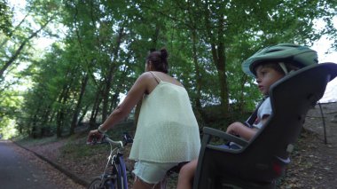 Mother riding bicycle with child in bike seat on green pathway outdoors. Family and weekend activity trip