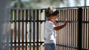 Çocuk balkon çitinin yanında dikilip ikinci kattan dışarı bakıyor. Tahta parmaklıklarda bekleyen çocuk dışarıya bakıyor.
