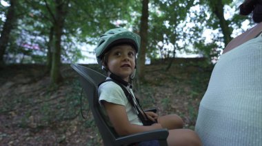 Mother riding bicycle with child in bike seat on green pathway outdoors. Family and weekend activity trip