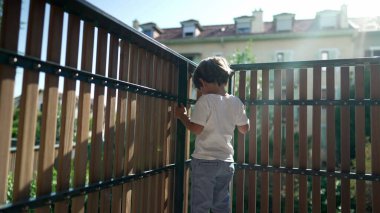 Balkon çitinin yanında duran küçük bir çocuk ikinci kattan dışarı bakıyor. Tahta parmaklıklarda bekleyen çocuk dışarı bakıyor.
