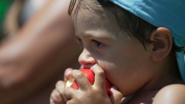 Küçük çocuk yaz günü dışarıda elma meyvesinden bir ısırık alıyor. Düşünceli şirin küçük çocuk dışarıda güneş ışığında sağlıklı şeyler yiyor.