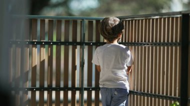 Pensive Little boy holding into balcony wooden fence at apartment building. Concept of security and protection of little boy at residential terrace