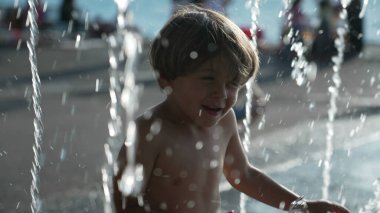 Küçük mutlu bir çocuk yaz tatillerinde açık hava parkında su jetlerine doğru koşuyor.