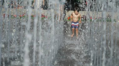 Küçük çocuk yaz günü şehir parkında su jetlerinde koşuyor. Küçük çocuk ferahlatıcı suda koşuyor yavaş çekimde sıçrıyor