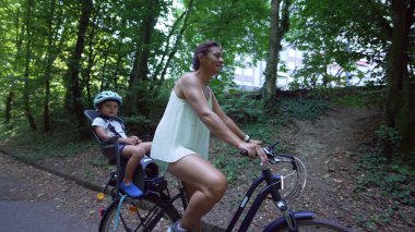 Mother riding bicycle with child in bike seat on green pathway outdoors. Family and weekend activity trip
