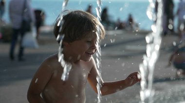 Küçük mutlu bir çocuk yaz tatillerinde açık hava parkında su jetlerine doğru koşuyor.