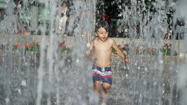 stock image Little boy running at water jets in city park during summer day. Small child runs in refreshing water splashing in slow motion