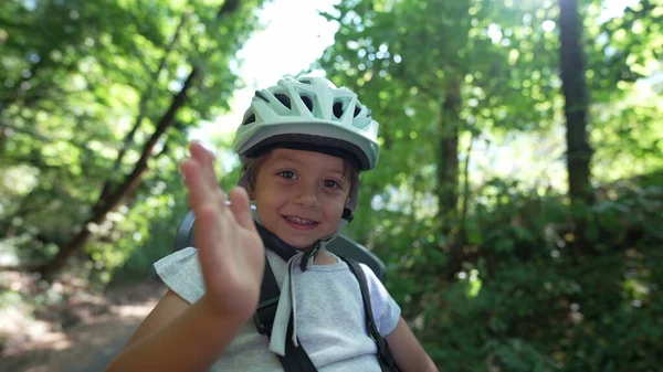 One happy child waving hello seated in bike seat wearing helmet protection. Adorable kid waves with hand in bicycle chair