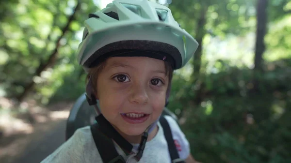 Happy child wearing bicycle helmet outdoors in forest park. Little boy weekend activity closeup portrait smiling seated in back of bike. Safety and protection concept