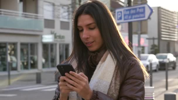 快乐的年轻女子微笑着拿着智能手机在城市街道上 20多岁的成年女子喜欢上网看城市人行道上的电话 — 图库视频影像
