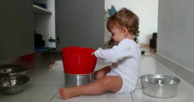 Cute baby on kitchen floor playing with kitchen utensils, playful one year old toddler boy holding pots and pans