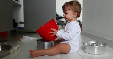 Cute baby on kitchen floor playing with kitchen utensils, playful one year old toddler boy holding pots and pans