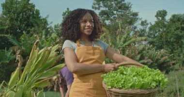 Güneşli bir günde, elinde taze organik salata dolu bir sepetle bir çiftlikte duran genç bir Afrikalı Amerikalı kadın çiftçinin portresi. Kadın çiftçi iş üniforması giyiyor ve yüzünde mutlu bir ifade var..