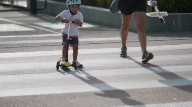 Scooter 'lı, koruyucu kasklı bir çocuk karşıdan karşıya geçiyor. Anne ve çocuk yaya geçidi zebra hatlarında.