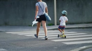 Anne ve çocuk karşıdan karşıya kask takarak ve scooter 'la geçiyorlar. Şehrin yaya geçidi zebra hatlarında aktif küçük bir çocuk.