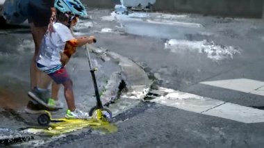 Anne ve çocuk yaya geçidi zebra hatlarında karşıdan karşıya geçiyorlar. Küçük çocuğu koruyan ebeveyn şehir sokaklarında kasklı scooter kullanıyor.