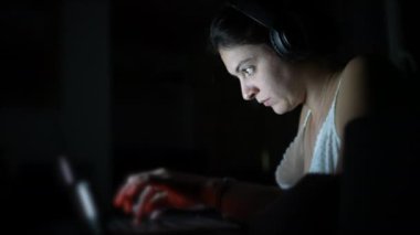 Focused woman typing on laptop keyboard in the dark wearing headphones. Person working in the dark. Modern lifestyle