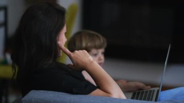 Anne, dizüstü bilgisayarın önünde çocuğuyla evde çalışıyor. Çocuk bebekle kanepede oturmuş bilgisayara bakıyor.