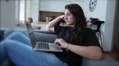 Woman browsing internet with laptop computer sitting on couch at home. Person looking at screen. Modern lifestyle