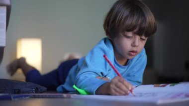 Odaklanmış çocuk kırmızı renkli kalemle kağıda çiziyor. Evde yaratıcı aktiviteler yapan konsantre bir çocuk yerde yatıyor.