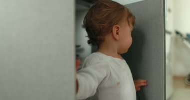 One year old baby boy opening kitchen cabinet cupboard closer door. Infant toddler standing at kitchen floor