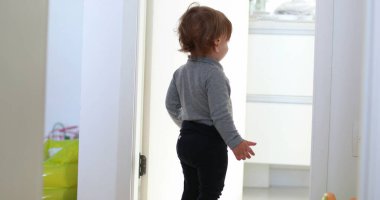 One year old baby toddler standing observing, infant child stands by bathroom door