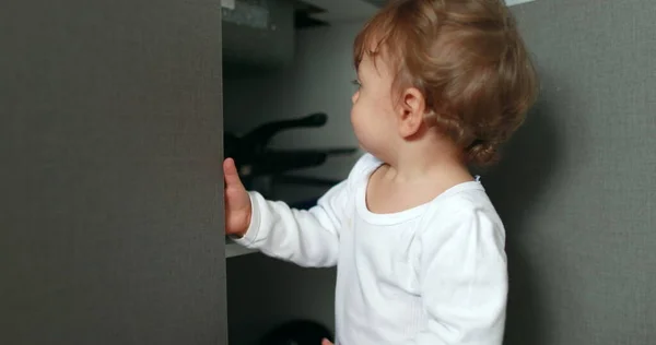 One year old baby boy opening kitchen cabinet cupboard closer door. Infant toddler standing at kitchen floor