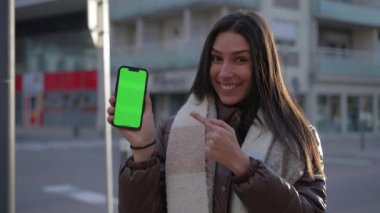 Şehir sokağındaki krom renkli yeşil ekranla akıllı telefondan video içeriğini paylaşan neşeli genç bir kadın. Mobil ekranı kameraya gösteren ve parmağıyla ekranı gösteren bir kadın.