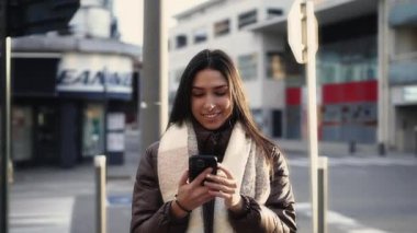 Şehir caddesinde dururken cep telefonu ekranına bakan mutlu, genç bir esmer kadın.