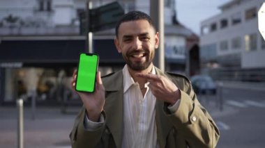 Orta Doğulu mutlu bir insanın portresi telefon ekranında kroma tuşlu yeşil ekranla içeriğini gösteriyor. Sokakta durup ekranı işaret eden ve gülümseyen bir adam. 