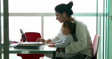 Mother multi-tasking, holding baby infant and using computer laptop at home. Candid authentic and real life mom working and parenting