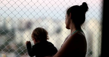 Mother holding baby standing by balcony window looking outside