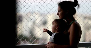 Pensive mother holding baby by balcony window looking outside