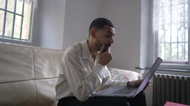 ラップトップコンピュータの前でソファに座っている間にオンラインで成功したメッセージに積極的に反応する幸せな若い中東の男が興奮しました — ストック動画
