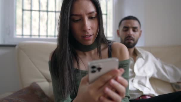 年轻女子坐在男友旁边的沙发上 在智能手机装置上读信息 有全神贯注表情的成年女孩 — 图库视频影像