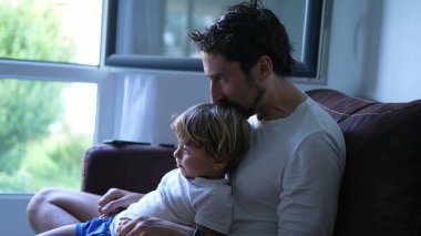 Evdeki kanepede baba kucağında oturan oğlum. Baba ve çocuğun birbirine bağlandığı otantik gerçek bir aile hayatı. Ebeveyn çocuğu sevgi ve sevgiyle öpüyor.