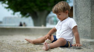 Küçük bir çocuk kum havuzunda oturmuş, kürek ve kumla oynuyor. Çocuk yaz günü dışarıda oynuyor.