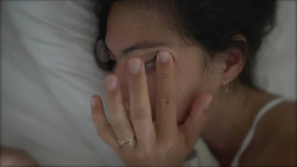 Woman rubbing eye laid in bed looking at phone. Tired sleepy female person rubs eye with finger