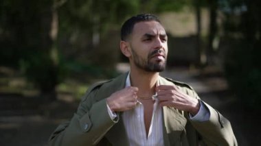 Güneşli bir günde parkın dışında yürüyen ve ceketini ayarlayan kendinden emin genç bir Arap. Modaya uygun mutlu insan.