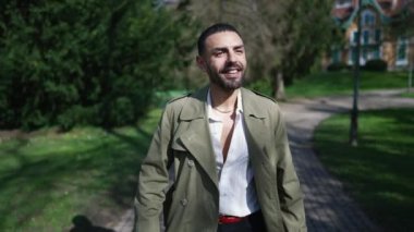 Neşeli genç bir Arap adam güneşli bir parkta güneşli bir şekilde yürüyor, güneşin sıcaklığında güneşleniyor ve dışarıdaki güzelliğin tadını çıkarıyor.