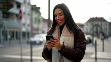 Mutlu bir genç kadın elinde akıllı telefon cihazıyla şehrin dışında yürürken gülüyor ve gülümsüyor.