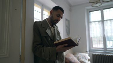 Duvara yaslanmış kitap okuyan bir adam. İnsan evde konsantre olup ders çalışır.