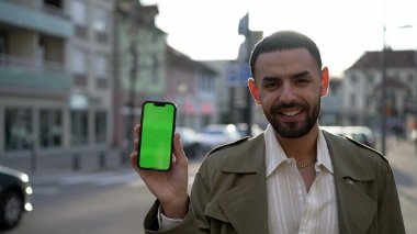 Neşeli bir Faslı genç adam krom yeşil ekran anahtarıyla telefon ekranını kameraya gösteriyor. Şehir kaldırımında gülümsüyor.