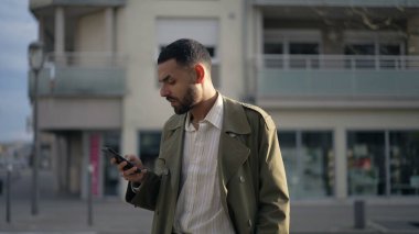 Ortadoğulu genç bir adam şehir caddesinde duruyor ve telefonları kontrol ediyor. Çevrimiçi içeriğe bakan bir Arap erkek.