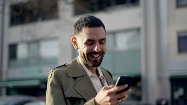 Orta Doğulu mutlu bir adam şehir caddesinde güneş ışığıyla beklerken telefon kullanıyor. İnternette sosyal medya içeriğine gülen ve gülümseyen Faslı bir erkek.